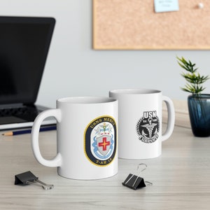 May include: Two white ceramic mugs with a navy blue and gold design. One mug features the USNS Mercy logo with the text "USNS MERCY" and "T-AH 19". The other mug features the US Navy Medical Corps logo with the text "USN" and "CORPSMEN".