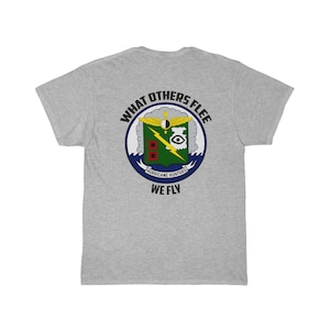 May include: A pale grey t-shirt with a circular emblem. The emblem has the text "WHAT OTHERS FLEE" at the top and "WE FLY" at the bottom. The emblem includes a lightning bolt, an eye, and the words "HURRICANE HUNTERS".