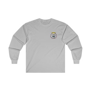 Peut inclure: Un t-shirt à manches longues gris clair avec un petit écusson brodé sur la poitrine gauche. L'écusson représente un blason avec un aigle ailé, un bouclier et le texte "V.F.W. POST 9088".