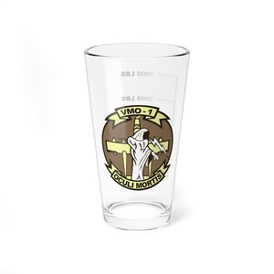 Op de afbeelding: Een helder glazen pintglas met een bruine en gele afbeelding van een squadroninsigne. Het insigne toont een schedel en gekruiste beenderen met de tekst "VMO-1" en "OCULI MORTIS".