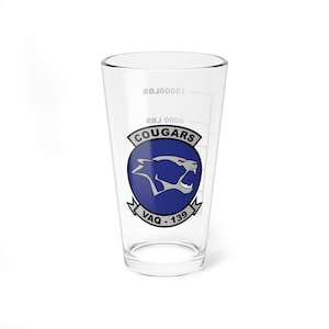 May include: A clear pint glass featuring the "Cougars" emblem, a blue circle with a cougar silhouette and the text "VAQ-139". The glass has measurement markings for 8000 lbs and 12000 lbs.