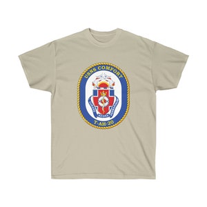 May include: A beige t-shirt with a blue and gold USNS COMFORT T-AH-20 crest. The crest features a snake wrapped around a staff with a cross at the top. The words "CURARE AEGRA PERMARINUM" are written around the snake.