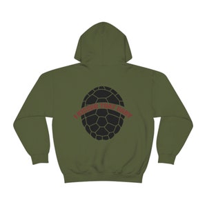 Peut inclure: Sweat à capuche vert olive avec un motif de carapace de tortue noire au dos. Le texte "I KISSED THAT BABY" est imprimé en rouge au-dessus de la carapace. Le sweat à capuche a une capuche et des manches longues.