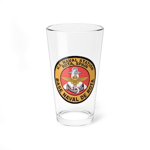 May include: A clear glass pint glass with a round, yellow and black sticker featuring the text "U.S. NAVAL STATION ROTA, SPAIN BASE NAVAL DE ROTA" and a red and white anchor with wings.
