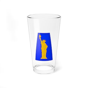 Puede incluir: Un vaso de pinta transparente con un diseño azul y dorado de la Estatua de la Libertad. El diseño es rectangular y está centrado en el vaso. El vaso está vacío y sobre una superficie blanca.