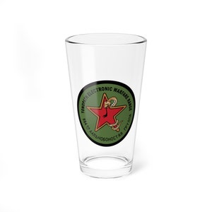 May include: A clear glass pint glass with a green and black circle graphic. The graphic features a red star with a snake wrapped around it. The text around the graphic reads "TONOPATH ELECTRONIC WARFARE RANGE 554 ВВ АЛЬБОНОСТЬ ЯВ ТРУДЛЯ".