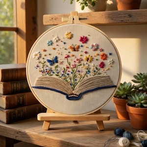 Könnte beinhalten: Gesticktes Kunstwerk in einem Stickrahmen, das ein offenes Buch mit bunten Blumen und Schmetterlingen zeigt. Das Kunstwerk wird auf einer Holzstaffelei präsentiert. Das Design umfasst Farben wie Blau, Rosa, Gelb und Orange.