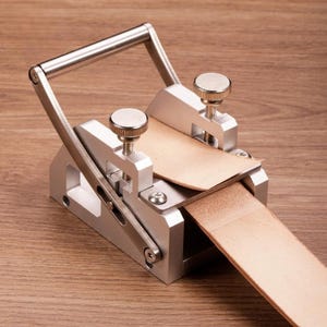 Pode incluir: Uma ferramenta de metal prateado para raspar couro com um cabo articulado, dois botões serrilhados ajustáveis para controle de tensão e uma base plana. Uma tira de couro bege claro está inserida no mecanismo, passando pela guia metálica.
