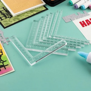 May include: Clear acrylic rulers and angle guides are arranged on a teal surface. The rulers have inch and fractional inch markings. Craft supplies, including markers and paper, are in the background. The word "HA" is visible on a white card.