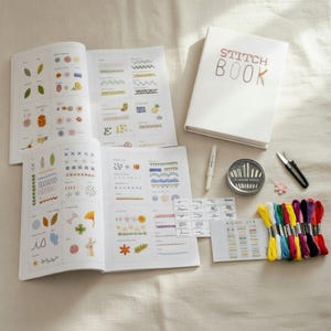 May include: A white "Stitch Book" with gold lettering is next to open instructional books with colourful stitch examples. A needle case, scissors, pen, and embroidery floss in various colours complete the craft kit.