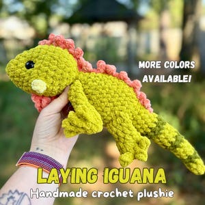 May include: A lime green crochet plushie in the shape of an iguana with a textured surface, a light yellow snout, and a row of pink spikes along its back. The text on the image reads: MORE COLORS AVAILABLE! LAYING IGUANA Handmade crochet plushie.