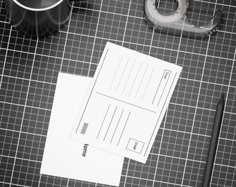 May include: Overhead shot of a workspace with a black mug, tape dispenser, pencil, and stationery. Two white postcards and a white envelope are on a black grid surface. The postcards have lines for writing and a stamp box.