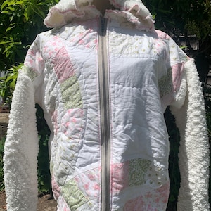 May include: A zip-up hooded jacket constructed from a patchwork of white, pink, and sage green floral fabric. The garment features long sleeves made of white textured sherpa fleece, a matching sherpa-lined hood, and a white lace trim at the hem.