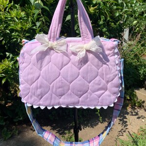 May include: A light purple quilted handbag featuring two top handles and a long, adjustable shoulder strap with a blue, pink, and yellow plaid pattern. The front is decorated with two white lace bows and a white scalloped trim along the bottom edge.