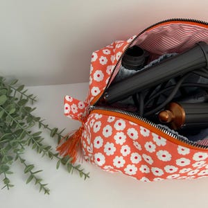 Könnte beinhalten: Eine offene Kosmetiktasche mit orangefarbenem Blumenmuster, schwarzem Reißverschluss und orangefarbenem Quastenanhänger. Die Tasche enthält ein schwarzes und kupferfarbenes Haarstyling-Gerät. Das Innenfutter ist rot und weiß gestreift.