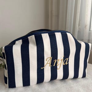 Può includere: Un beauty case a righe blu navy e bianche con chiusura a zip. Il nome "Anja" è ricamato in oro sul davanti. La borsa rettangolare è in tessuto.