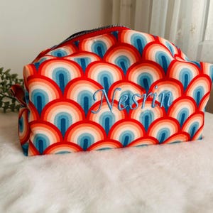 May include: A cosmetic bag with a vibrant retro rainbow pattern in red, orange, and blue. The bag has a red zipper and the name "Nesrin" embroidered in blue script. The bag is on a white surface.
