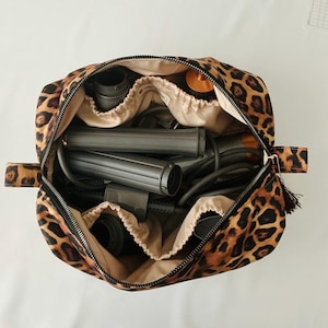 May include: Open leopard print cosmetic bag with a black zipper, containing hair styling tools. The bag has multiple compartments and a beige interior. The tools include a hair dryer, curling iron, and brush.