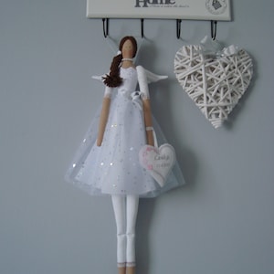 Può includere: Bambola angelo fatta a mano in un abito bianco con gonna di tulle e ali. La bambola ha i capelli castani e tiene in mano un cartello a forma di cuore. Sono visibili anche un cuore bianco intrecciato e un portachiavi a muro con la scritta "Sweet Home".