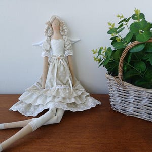 May include: A handcrafted fabric doll featuring blonde braided hair, white wings, and a beige tiered dress with a floral pattern. The doll has long limbs, white lace trim on the sleeves and hem, and a small button detail at the waistline.
