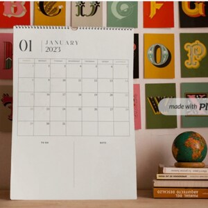 May include: A white calendar with a black border and the month of January 2023 printed on it. The calendar is hanging on a wall with colorful letters behind it.