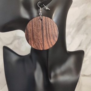May include: A circular earring featuring a dark brown wood grain pattern with visible vertical textures. The earring is suspended from a silver-toned metal hook wire, which connects to the top of the round wooden disc through a small metal jump ring.