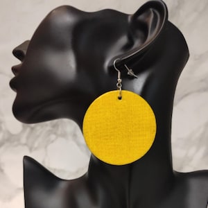 May include: A large, circular, bright yellow earring with a visible fabric-like texture. The earring features a silver-toned metal hook and a small jump ring connector, hanging from a black display bust to showcase its round shape and vibrant color.