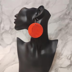May include: A pair of large, round orange earrings. The earrings are suspended from silver-colored hooks. The earrings are displayed on a black mannequin head against a marble-patterned background. The earrings are a bold, eye-catching accessory.