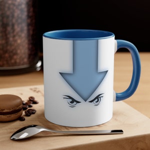 May include: White ceramic mug with a blue interior and handle. The mug features a light blue arrow design with stylized eyes. The mug is on a wooden surface with a spoon, coffee beans, and a pastry.