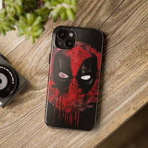 Puede incluir: Una funda de teléfono negra con un diseño de personaje de Deadpool rojo y negro. La funda tiene un aspecto desgastado de estilo grunge.