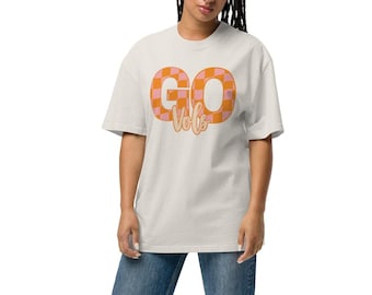 May include: A person wearing a cream-colored oversized t-shirt with the words "GO Vols" in orange and pink checkered letters. The person is also wearing blue jeans and white sneakers.