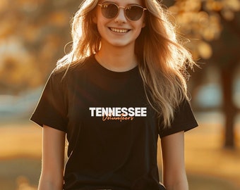 May include: A woman wearing sunglasses and a black t-shirt with the text "Tennessee Volunteers" in white and orange lettering. She is standing in a field with trees in the background.