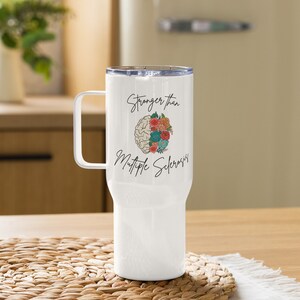 May include: White travel mug with a handle and a clear lid. The mug features the text "Stronger than Multiple Sclerosis" with a brain and floral design. The mug is designed to hold approximately 20 ounces.