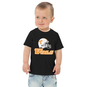 Peut inclure: T-shirt noir pour tout-petits avec un motif de casque de football blanc et orange et le mot "Vols" en orange. Le t-shirt est fait d'une matière douce, adaptée aux jeunes enfants.