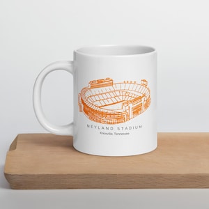 Peut inclure: Mug en céramique blanche avec une illustration orange du stade Neyland à Knoxville, Tennessee. Le design du stade est détaillé, avec le texte "NEYLAND STADIUM" et la ville et l'état imprimés en dessous.