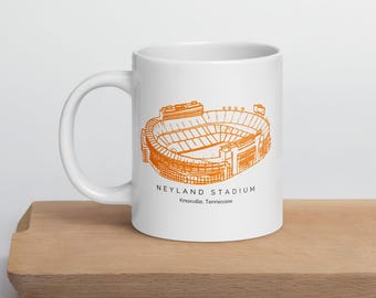 May include: White ceramic mug featuring an orange illustration of Neyland Stadium in Knoxville, Tennessee. The stadium design is detailed, with the text "NEYLAND STADIUM" and the city and state printed below the stadium image.