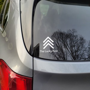 May include: A silver car with a white sticker on the back window. The sticker features a white arrow pointing upwards and the text "The Lucky Few".