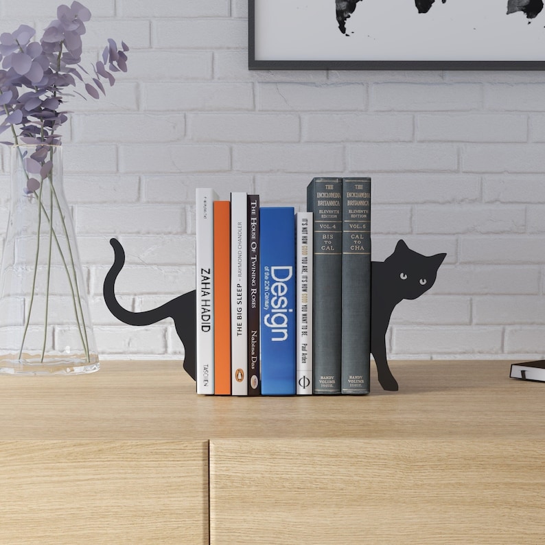 May include: Black metal cat bookends holding a row of books with titles like "Design", "The Big Sleep", and "The House of Twinning Roses".