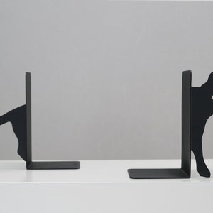 May include: A pair of black metal bookends shaped like cats. The cat on the left is looking to the right, while the cat on the right is looking to the left. Both cats are peeking out from behind the bookends.