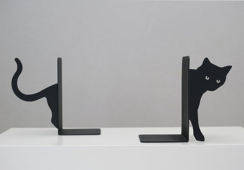 May include: A pair of black metal bookends shaped like cats. The cat on the left is looking to the right, while the cat on the right is looking to the left. Both cats are peeking out from behind the bookends.