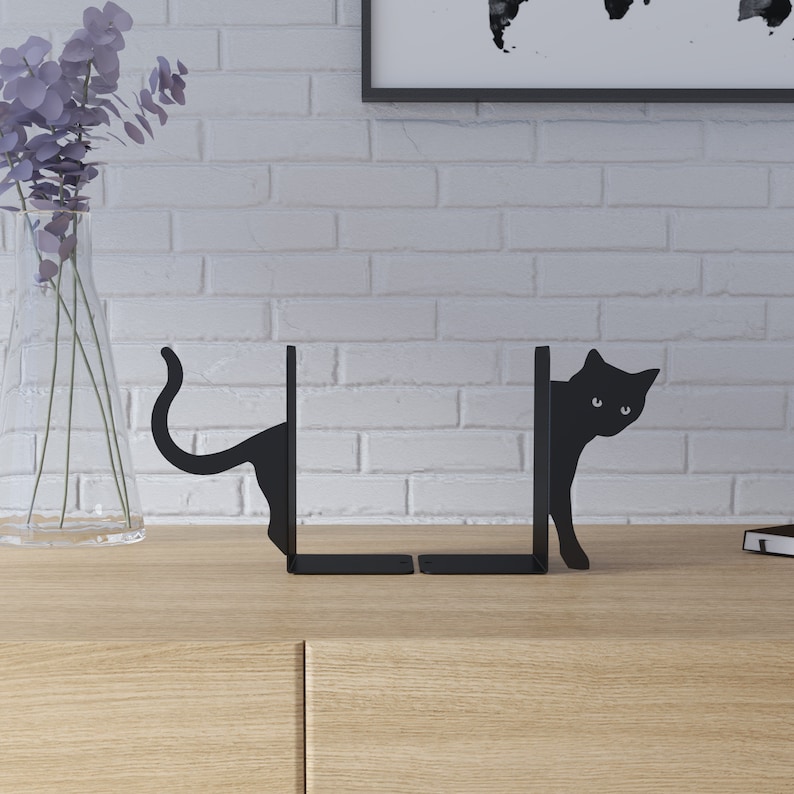 May include: Black metal bookends shaped like cats peeking around a corner. The bookends are a decorative way to hold books upright on a shelf.