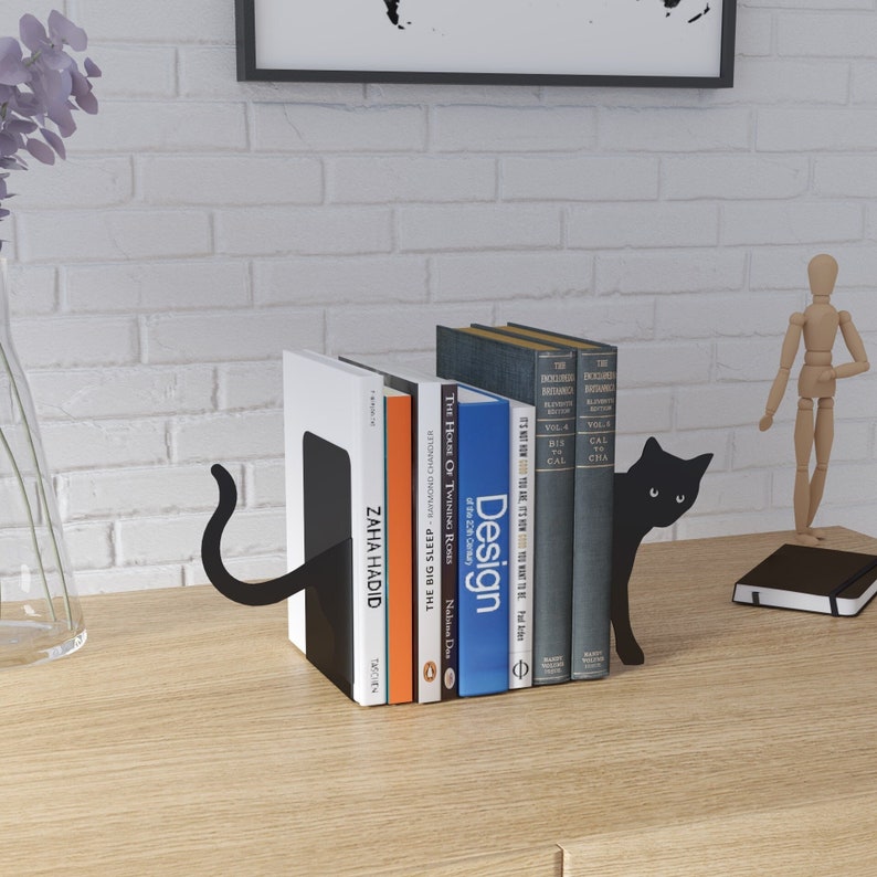 May include: Black metal bookends shaped like a cat with a tail curled around the back of the bookends. The bookends are holding up a stack of books with titles like "Design", "The House of Twining Roses", and "The Big Sleep".