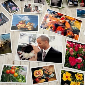 May include: A collection of various rectangular photographic prints scattered on a light wood surface. The images feature diverse subjects including a couple, koi fish, tulips, tomatoes, a golden statue, landscapes, and sunsets in various sizes.