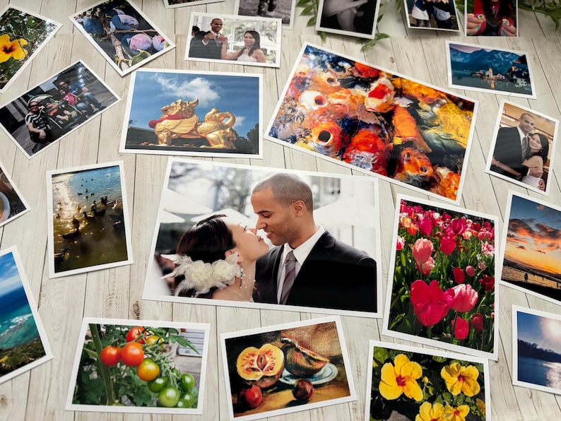 May include: A collection of various rectangular photographic prints scattered on a light wood surface. The images feature diverse subjects including a couple, koi fish, tulips, tomatoes, a golden statue, landscapes, and sunsets in various sizes.