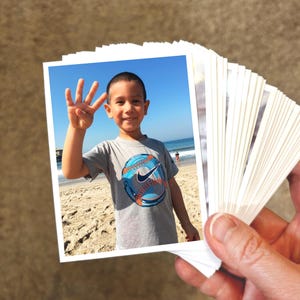 May include: A stack of small rectangular photographs with white borders. The top photo shows a child wearing a gray t-shirt with a blue and orange baseball graphic, holding up four fingers against a backdrop of a sandy beach and blue ocean.