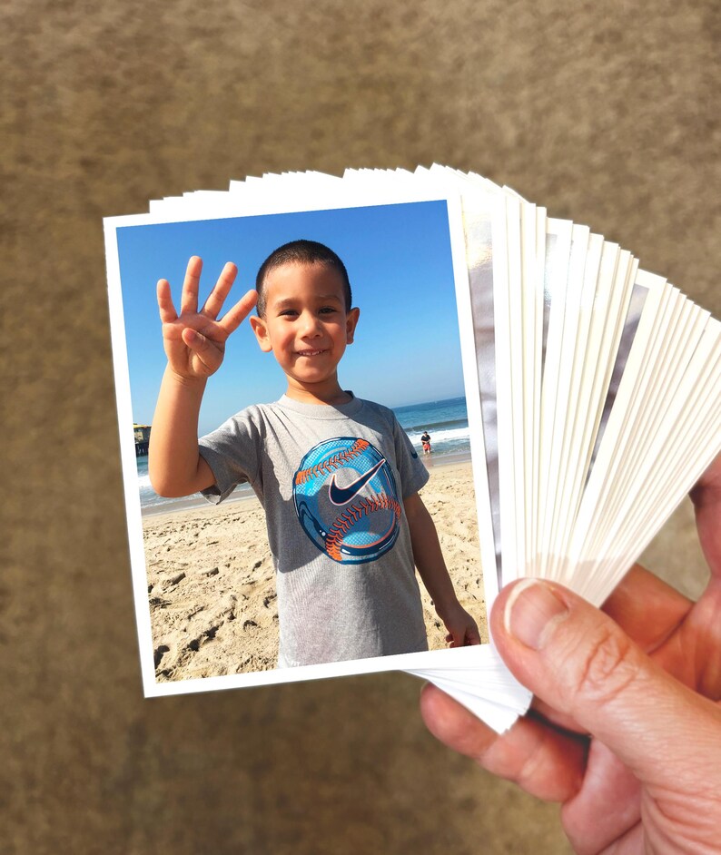 May include: A stack of small rectangular photographs with white borders. The top photo shows a child wearing a gray t-shirt with a blue and orange baseball graphic, holding up four fingers against a backdrop of a sandy beach and blue ocean.