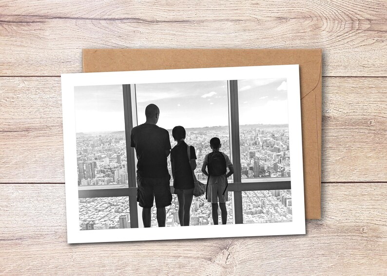May include: A black and white greeting card featuring a photograph of three individuals standing before a large window, looking out over a sprawling city skyline. The card is placed against a light brown envelope on a wooden surface.