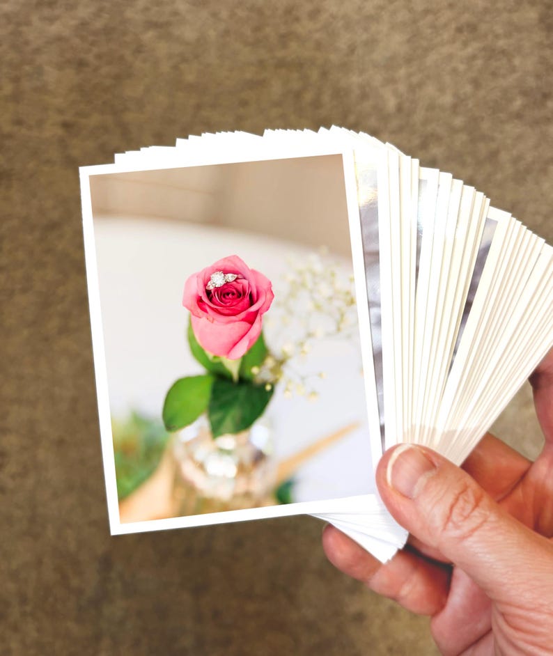 May include: A stack of rectangular photographic prints featuring a single pink rosebud with a silver ring resting inside the petals. The rose has green leaves and is accompanied by small white flowers in a clear glass vase, captured in a bright setting.