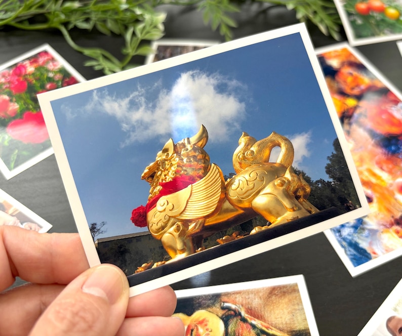 May include: A rectangular photograph featuring a shiny gold-colored mythical creature statue with wings and a red collar against a bright blue sky with a single white cloud. The print has a white border and is surrounded by other colorful photographs.