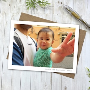 May include: A rectangular photo print featuring a baby in a green patterned shirt reaching toward the camera, held by a person in a white and blue shirt. The bottom right corner of the white border contains the text, Happy 1st Birthday, Emma!